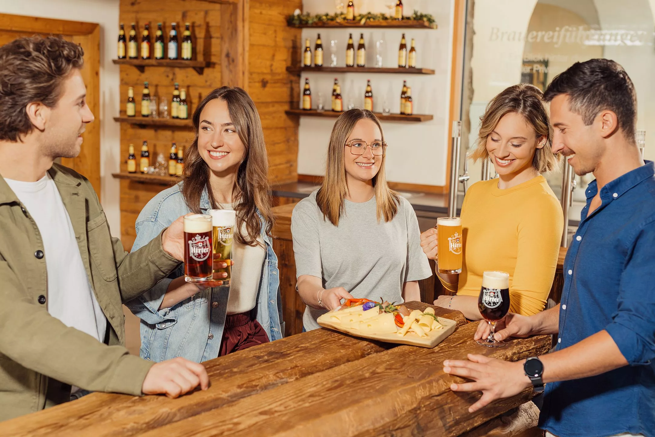 Fünf Personen stehen in der Hirter Bierathek um einen Holztisch auf dem eine Käseplatte steht und stoßen mit Hirter Bier an.