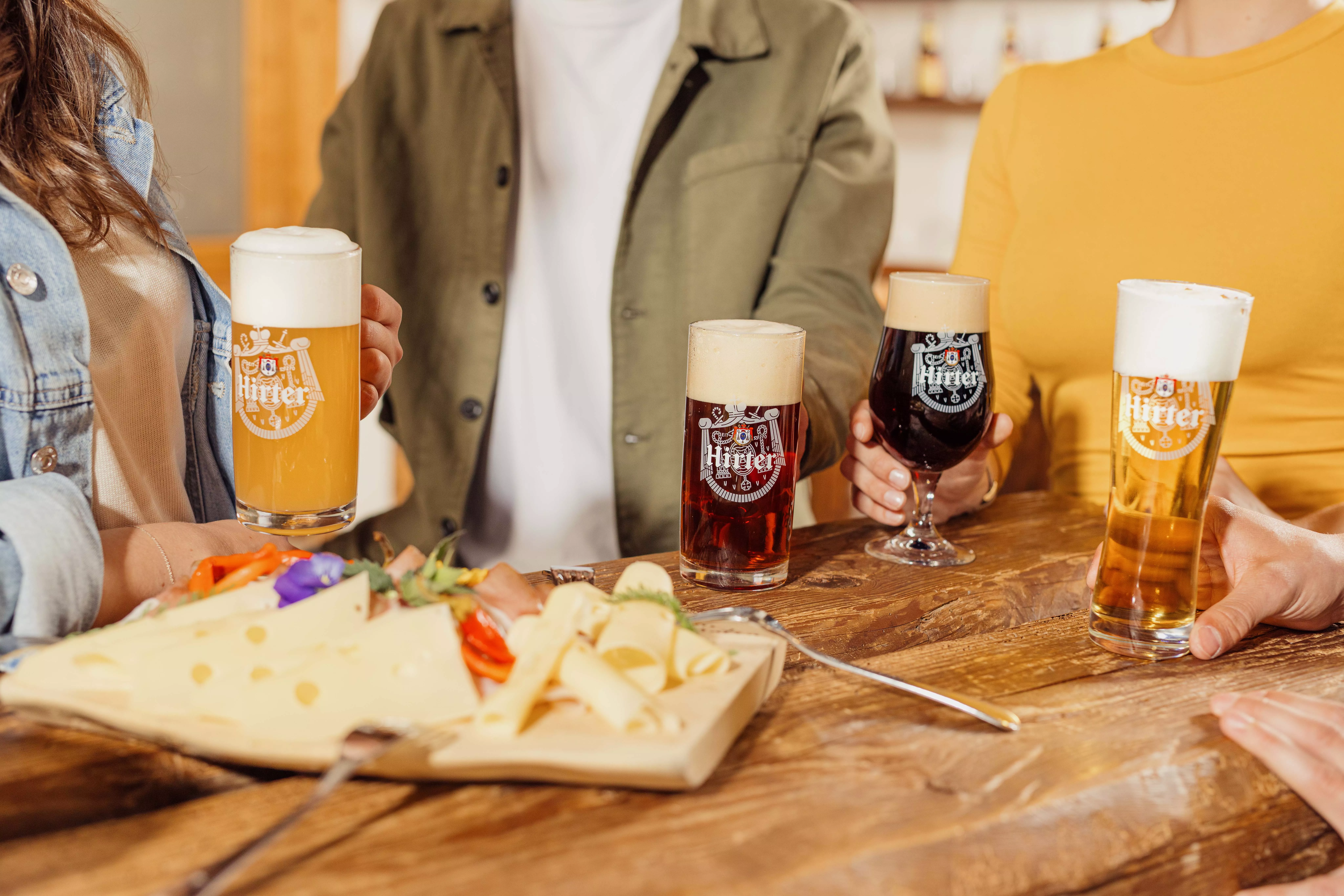Vier unterschiedliche Biere sind auf einem Tisch neben einer Käseplatte zu sehen mit Menschen im Hintergrund.