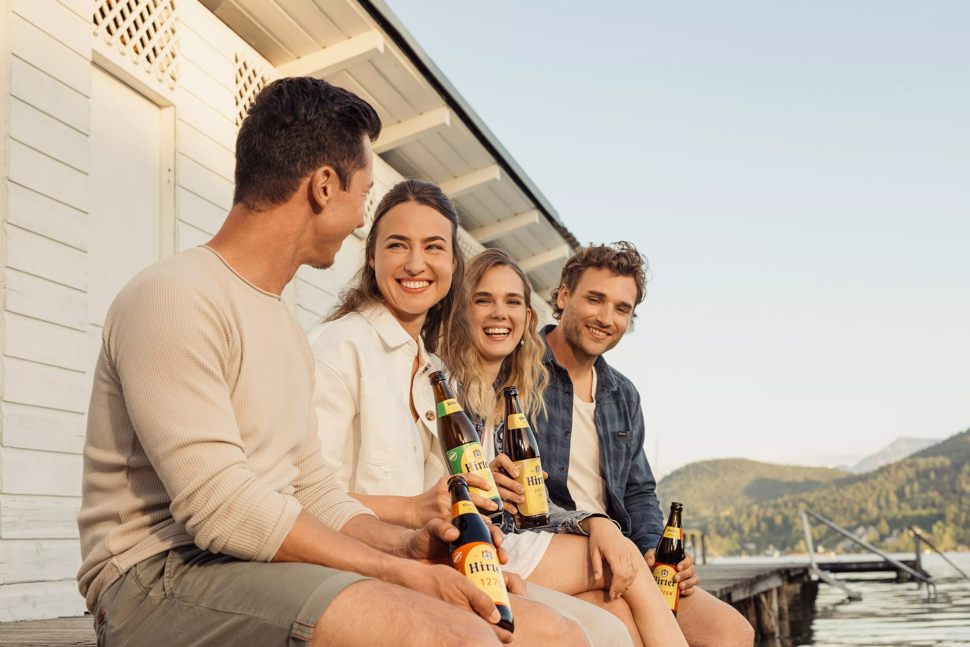 Vier Freunde sitzen an einem See an einem Steg und haben Hirter Bierflaschen.