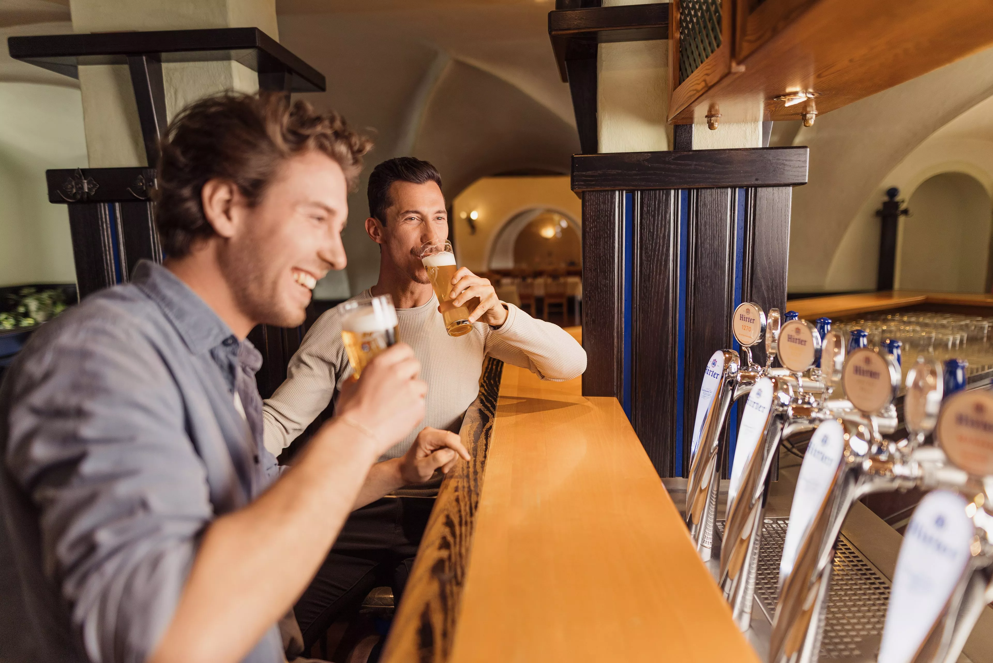 Zwei Männer sitzen an einer Schank. Beide lachen und haben Bier in der Hand.